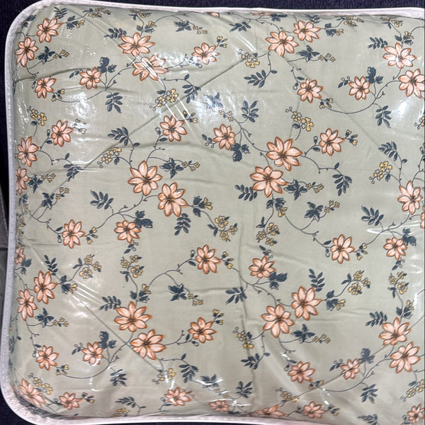 A square-shaped, shrink-wrapped mattress topper or quilted cushion with a floral pattern, shown on a dark blue carpet with a person's bare feet visible at the bottom edge.
