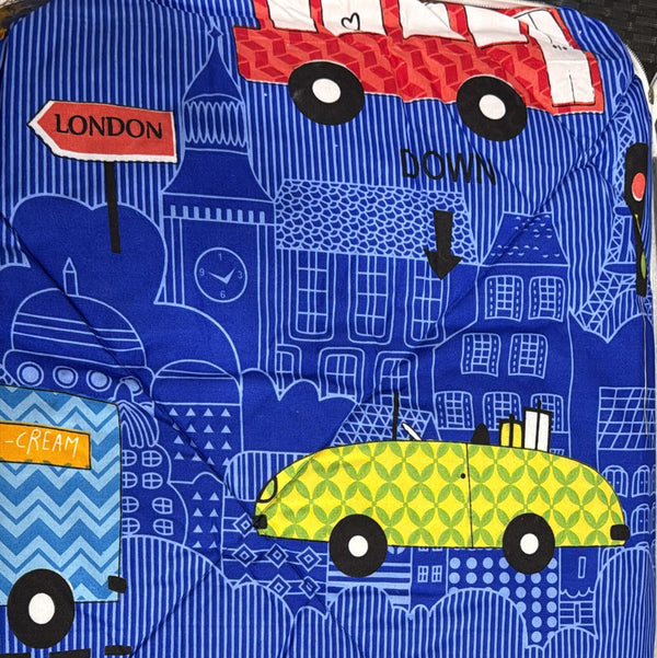 A blue quilted luggage cover featuring a London-themed print with illustrations of a red double-decker bus, a yellow taxi, a blue bus, and white line drawings of London landmarks and street signs.